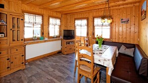 Interior de una cabaña de madera con una mesa de comedor, sillas, un sofá, un televisor y una zona de cocina.