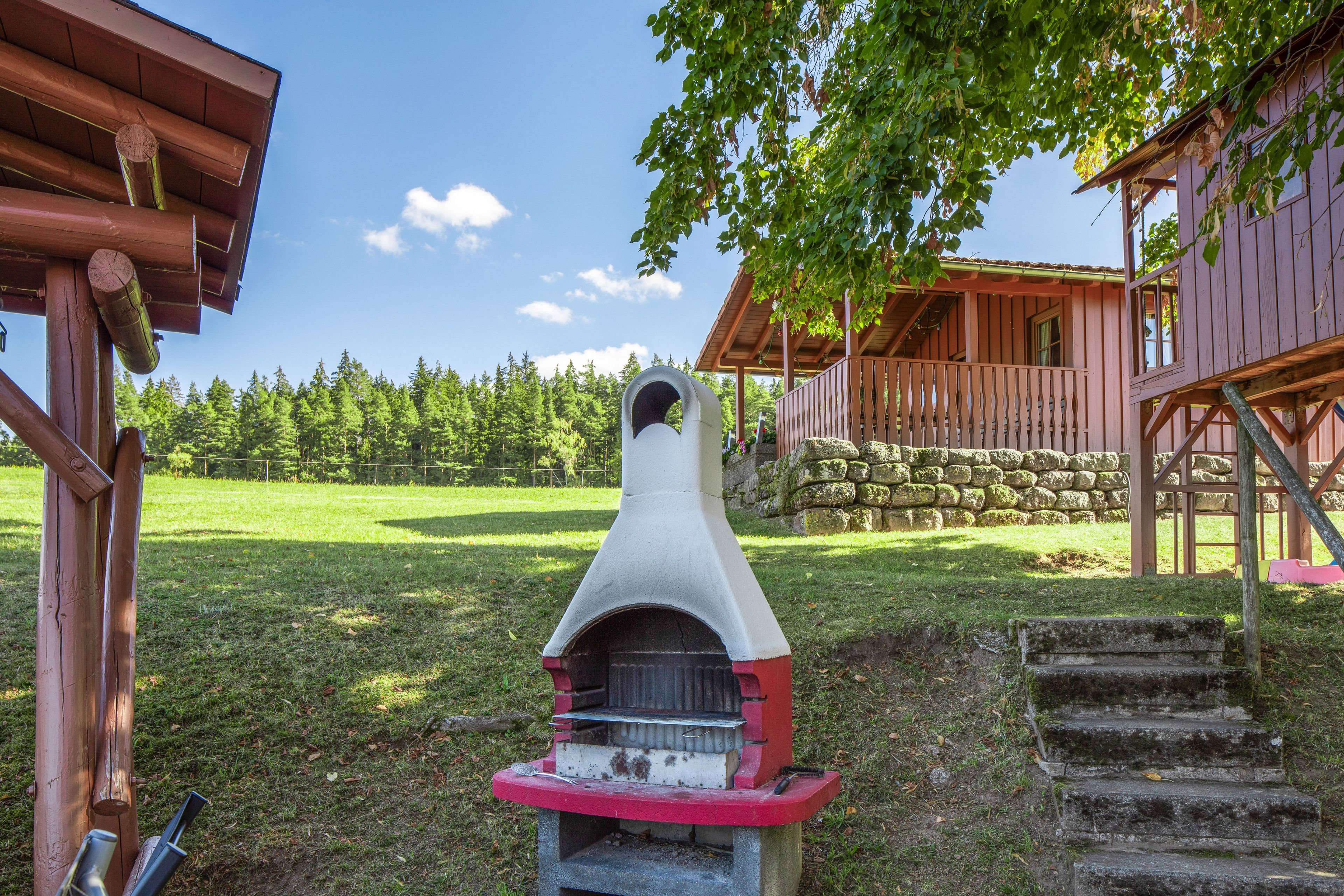 Uma área de churrasco rústica ao ar livre, com chaminé de pedra, degraus de madeira e um muro de pedra num campo verde e gramado.