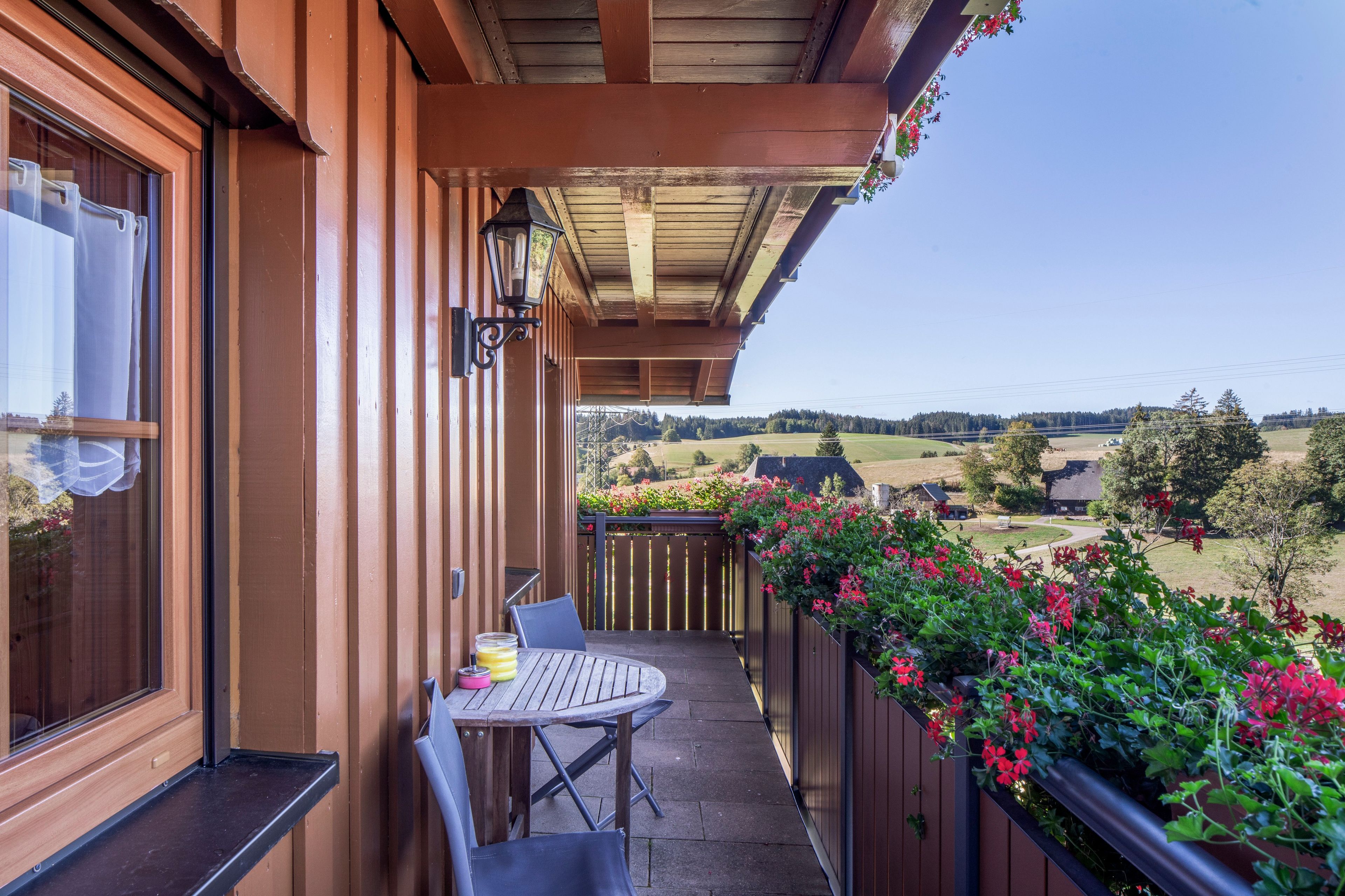 Ein Holzbalkon mit Tisch und Stühlen, mit Blick auf eine ländliche Landschaft mit Feldern und Häusern.