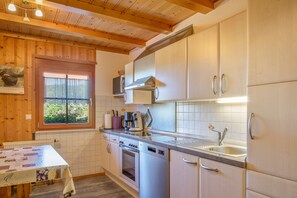A kitchen with wooden ceiling beams, a window with a view, a coffee machine, and stainless steel appliances. - Apartment 'Talaue' with Terrace, Barbecue Area, Playground and Countryside View (Sankt Georgen im Schwarzwald)