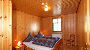 Una habitación de madera con cama, ventana, una silla y mesita de noche con lámpara.