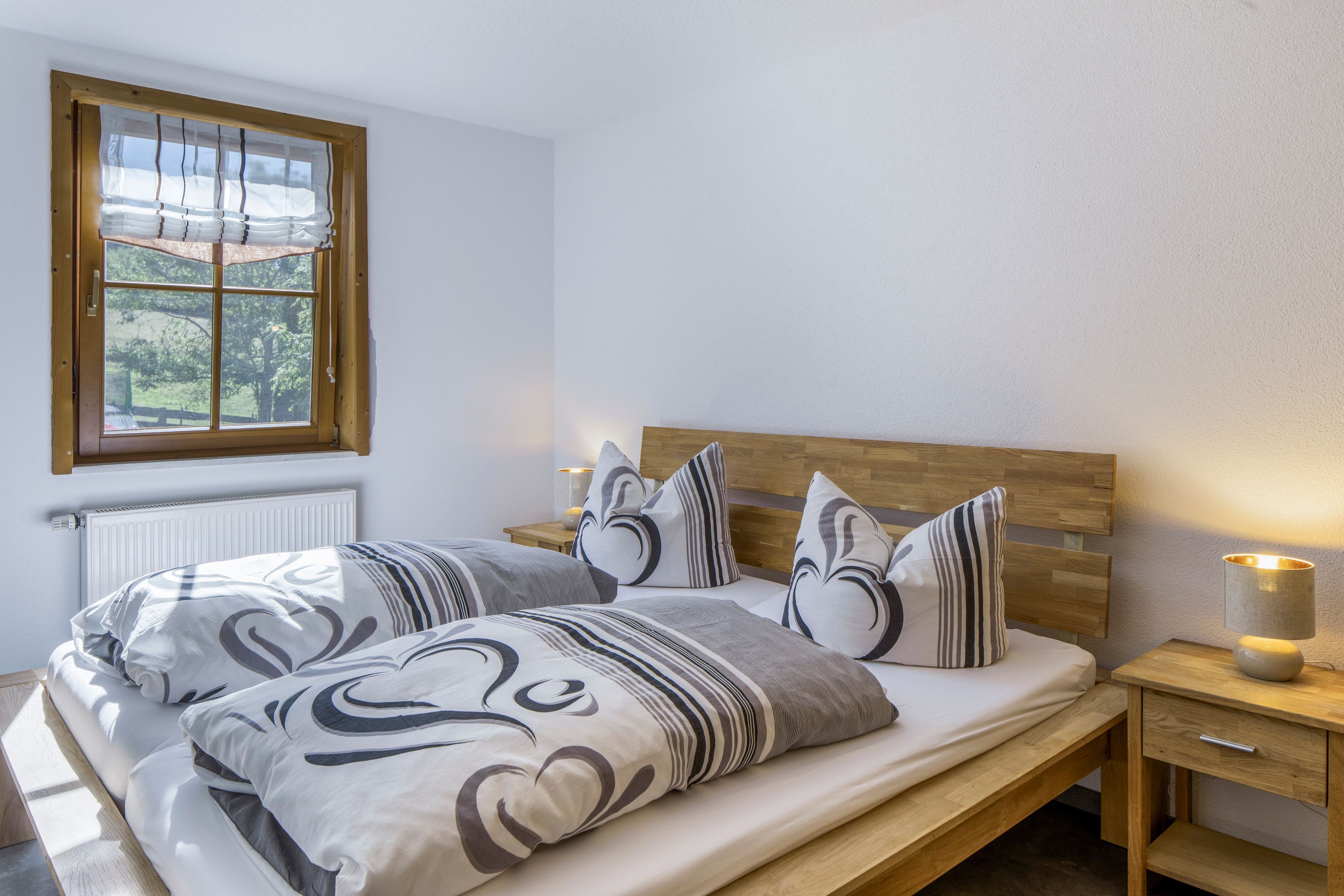A bedroom with a wooden headboard, a bed with patterned bedding, a bedside table with a lamp, and a window with curtains.