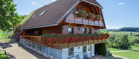 A two-story wooden chalet with a sloped roof, a balcony with red flowers, and a paved entrance area.