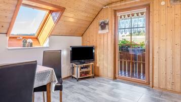 A cozy room with a sloped ceiling, a television on a wooden stand, a table with chairs, and a large window with a view of greenery outside.
