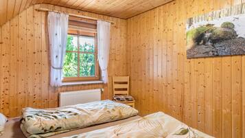 A wooden cabin bedroom with two beds, a radiator, a chair, and a window with curtains.