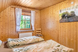 A wooden cabin bedroom with two beds, a radiator, a chair, and a window with curtains. - Apartment 'Waldblick' on the farm with terrace, barbecue area, playground and green views (Sankt Georgen im Schwarzwald)