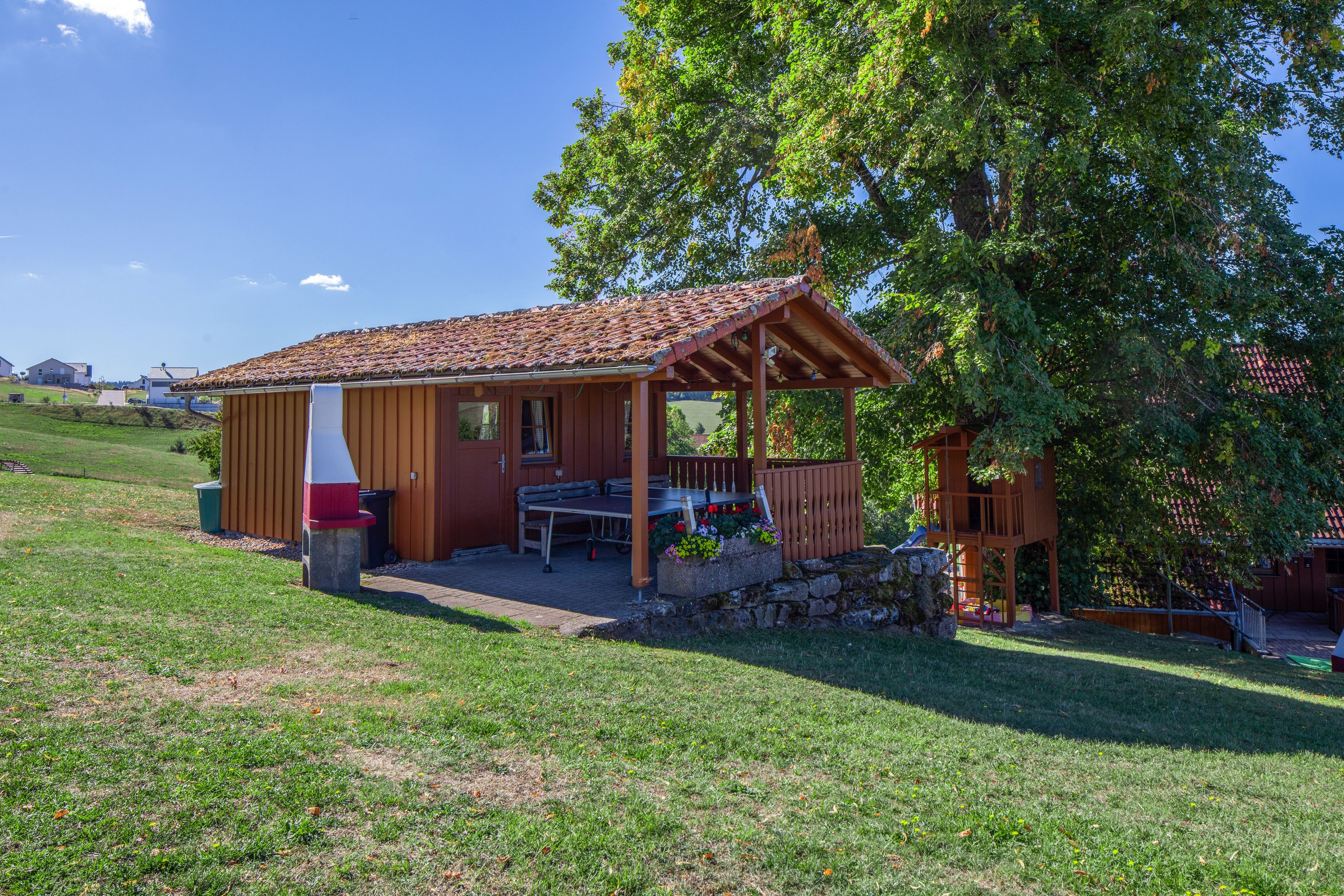 Una cabaña de madera con techo de tejas, un patio con mobiliario exterior y una pared de piedra.