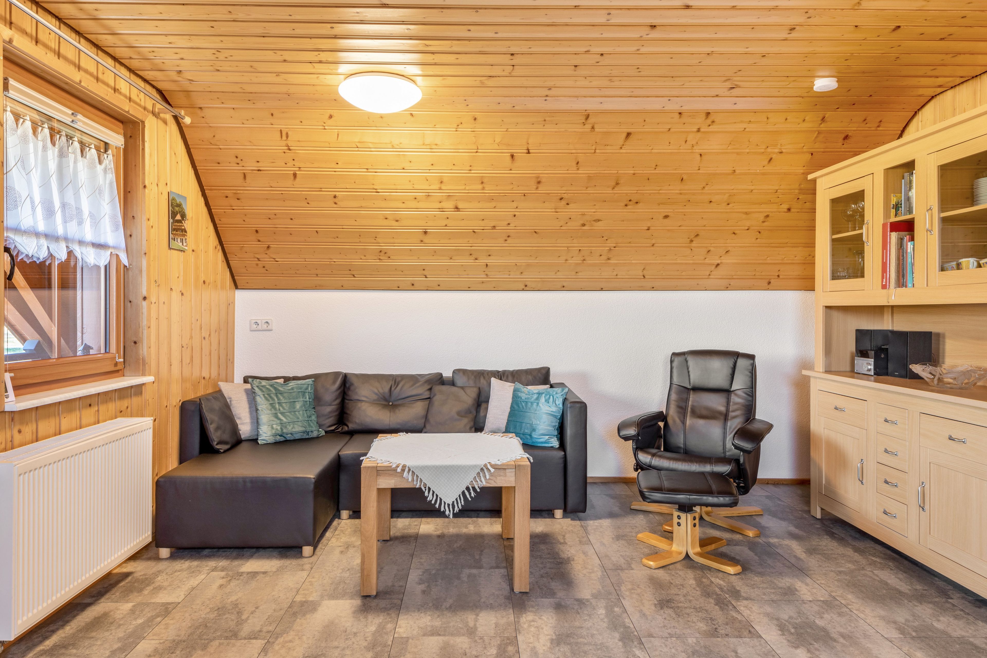 A cozy room with a wooden ceiling, a sofa, a chair, a table, and a cabinet with books and glassware.