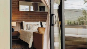 A bedroom with a brick wall, a bed with patterned pillows, a nightstand with a lamp, and a view through a glass door. - The Doeller House | Pool, Sauna & Speakeasy (Sedona)