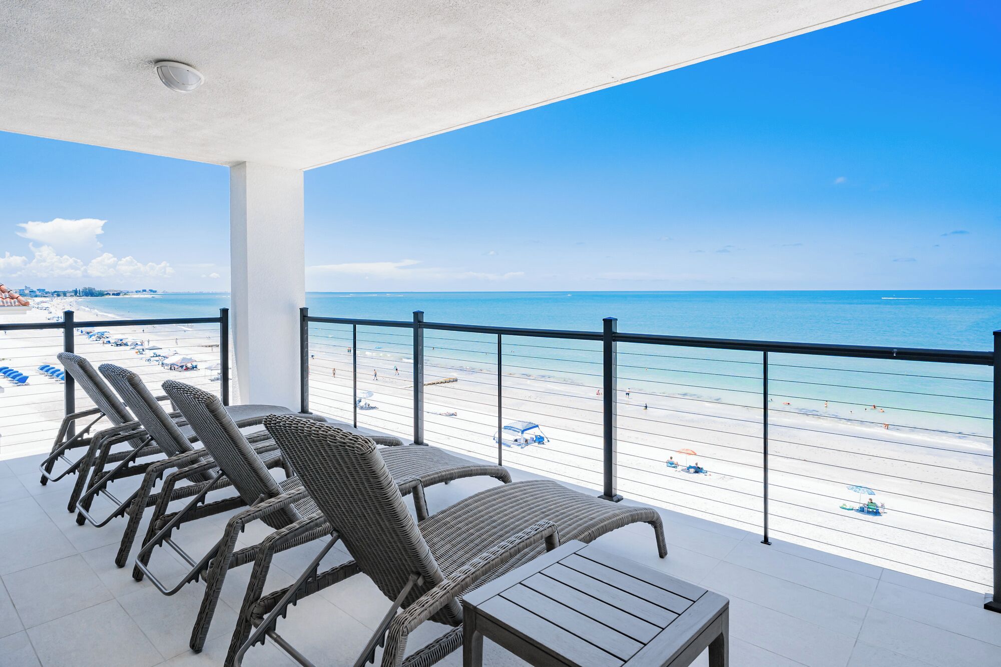 A balcony with wicker chairs and a table overlooking a beach with clear blue water.