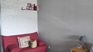 Quarto com sofá vermelho, mesa de cabeceira de madeira com abajur, tapete cinza e prateleira com livros e objetos decorativos.