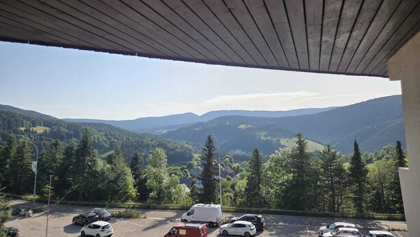 Um espaço de estacionamento com veículos, vista para colinas arborizadas e um edifício com telhado inclinado.