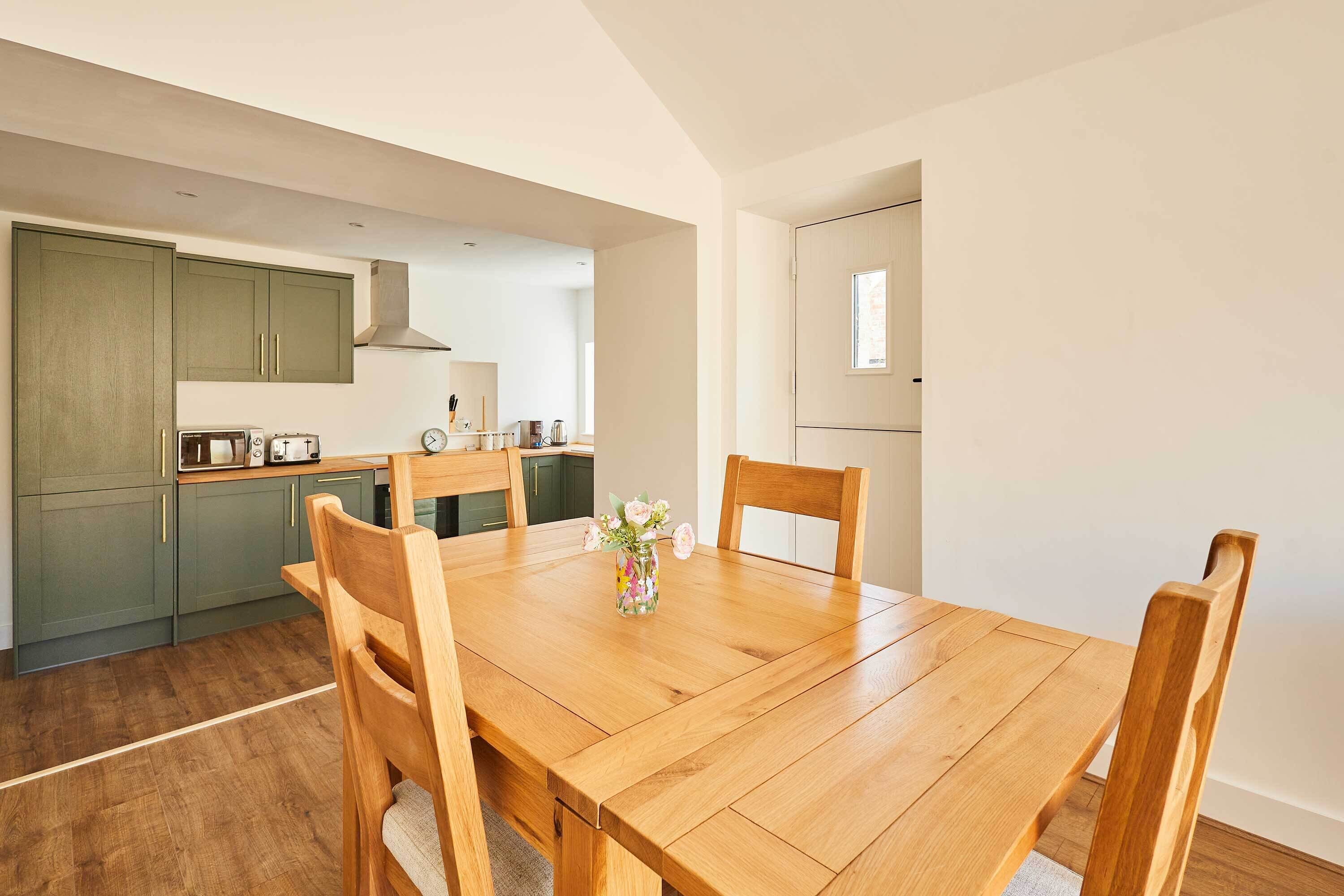 A dining area with a wooden table and chairs, a vase with flowers, and a kitchen in the background.
