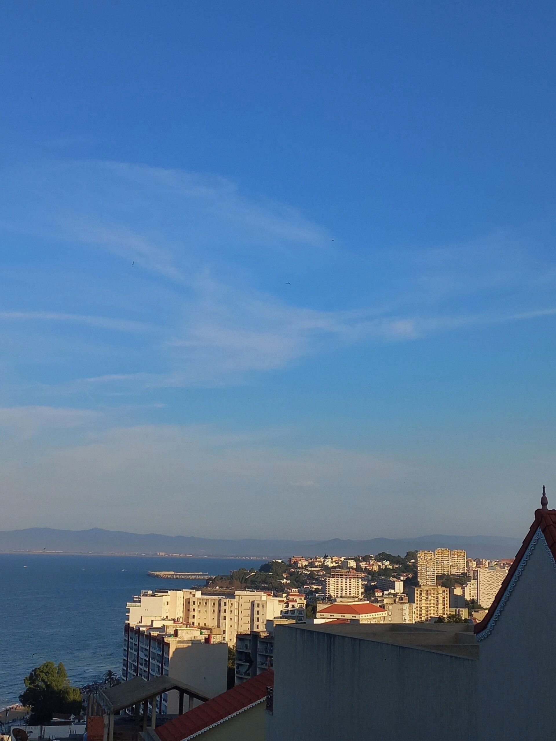 A coastal cityscape with buildings, a clear sky, and a view of the sea.