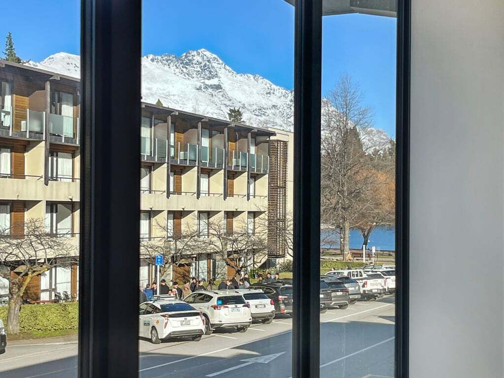 Ein mehrstöckiges Gebäude mit Parkplatz davor, einem blauen Schild und schneebedeckten Bergen im Hintergrund.