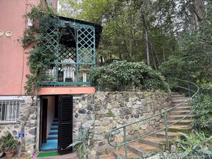 A stone staircase leading to a wooden door with a black shutter, surrounded by greenery and a pink building. - Il Pulcino della Pavona - sea view and garden (Monterosso al Mare)