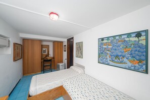 Ein Schlafzimmer mit einem Bett, einem Schreibtisch und einem Sessel. An der Wand hängt ein Gemälde und ein Klimagerät ist ebenfalls angebracht.