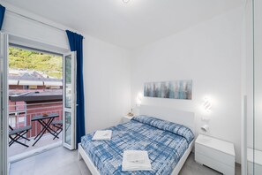 A bedroom with a bed, a nightstand, a wall-mounted lamp, a window with curtains, and a balcony with a table and chairs. - Charlie 89 Modern Apt with A/C in the center (Monterosso al Mare)