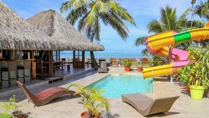 Un área de piscina con sillas de descanso, una estructura con techo de paja y un tobogán de colores.