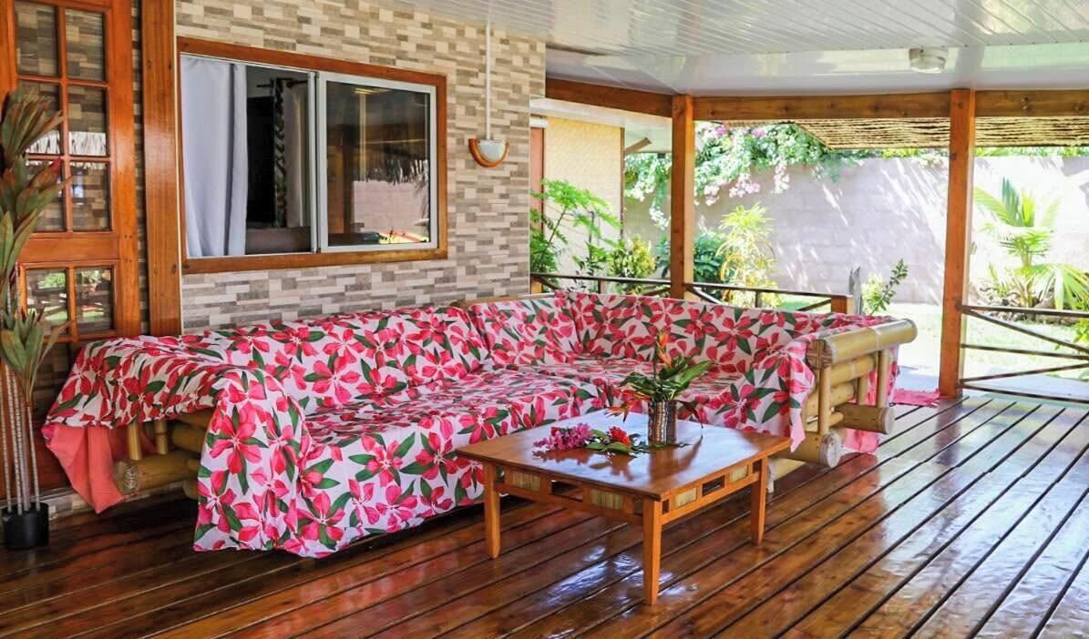A wooden deck with a sofa covered in a floral-patterned blanket, a wooden table with a vase, and a window with curtains.