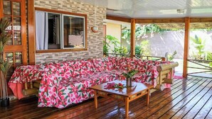 Una terraza de madera con un sofá cubierto por una manta estampada, una mesa de madera con un florero y una ventana con cortinas.