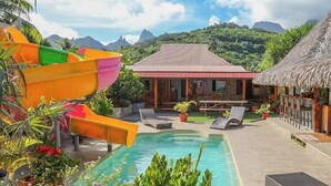 Un área de piscina con tobogán, sillas de descanso y un edificio con techo de paja.