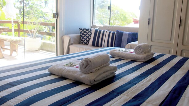 A bed with striped bedding, a window with a view of greenery, and a door to a balcony. - Modern loft 5 minutes from Geribá with a balcony, grill, and pool—pets welcome! (Armação dos Búzios)