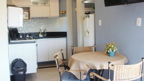 A compact kitchen with white cabinets, a black countertop, a dining table with wicker chairs, and a TV mounted on the wall. - Modern loft 5 minutes from Geribá with a balcony, grill, and pool—pets welcome! (Armação dos Búzios)
