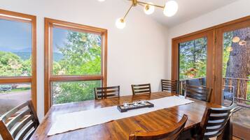 A dining area with a wooden table, six chairs, and a chandelier. Large windows offer views of greenery and a mountain.