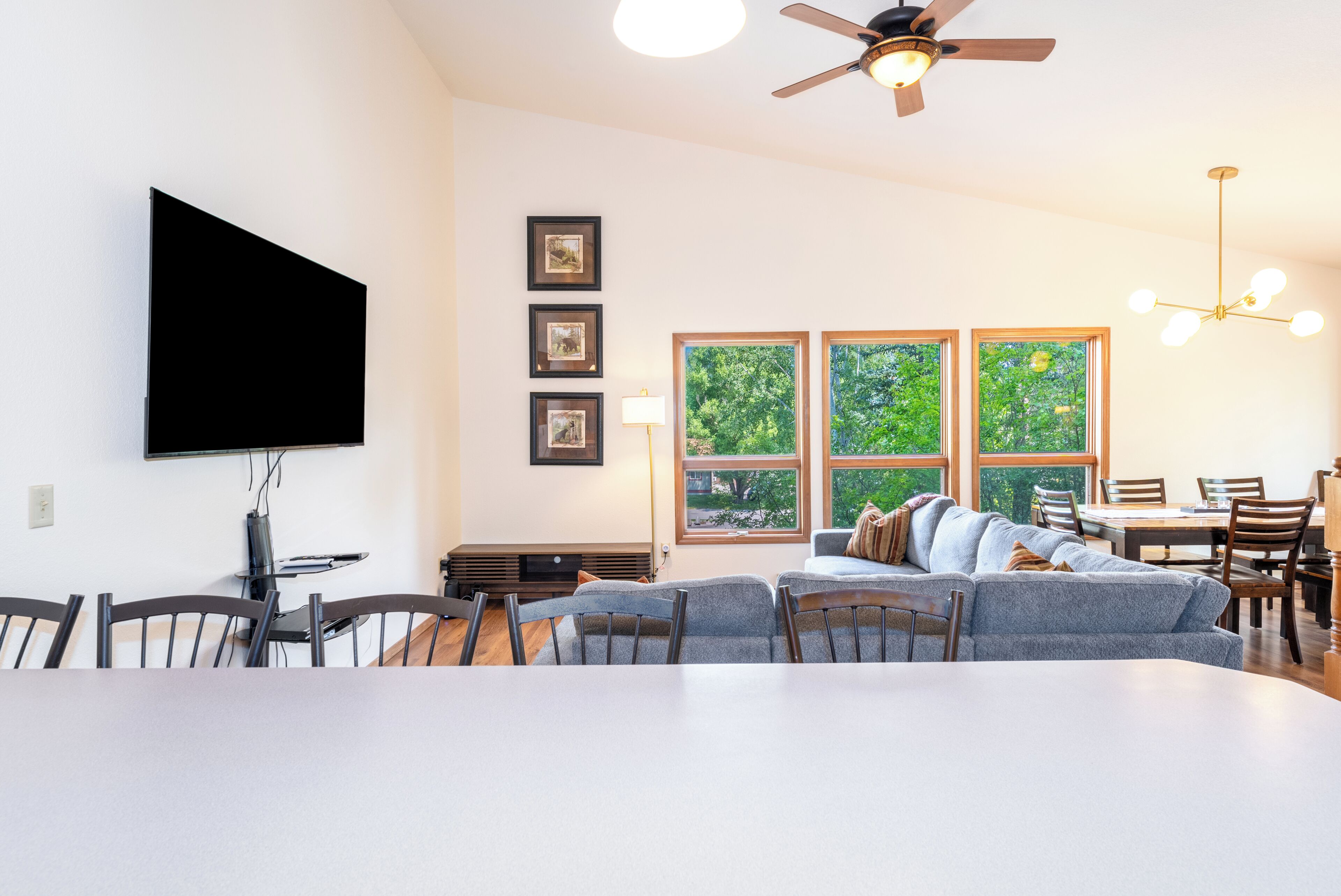A modern living room with a flat-screen TV, a ceiling fan, a dining area with a table and chairs, and a large window with a view of trees.