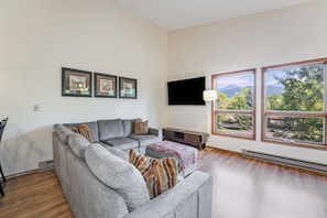 A living room with a grey sofa, a flat-screen TV, framed pictures, and large windows with a view of trees and mountains. - New Listing! Montana Ridgeline Retreat | GNP + Ski + Hike (Whitefish)
