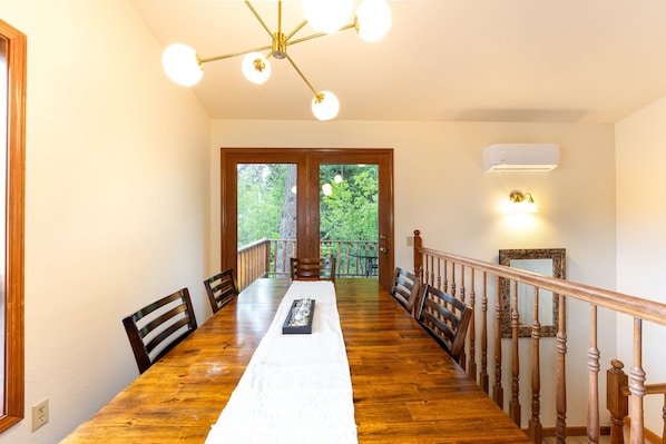 A dining room with a long wooden table, chairs, a chandelier, and a view through a door to an outdoor deck.