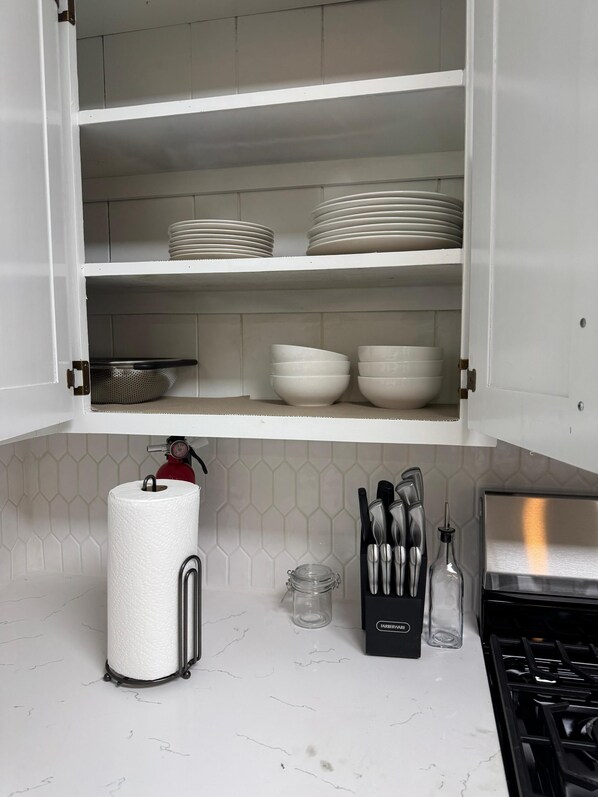 Una cocina con armarios blancos, una estufa a gas, un rollo de toallas de papel, un colador, un bloque de cuchillos y un frasco de vidrio.