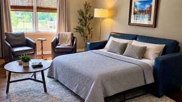 A bedroom with a bed, two armchairs, a side table, a lamp, a painting, and a window with curtains.