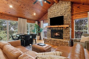 Ein gemütliches Wohnzimmer mit Kamin aus Stein, Ledersofas, einem Essbereich mit Tisch und Stühlen sowie großen Fenstern mit Blick ins Freie.