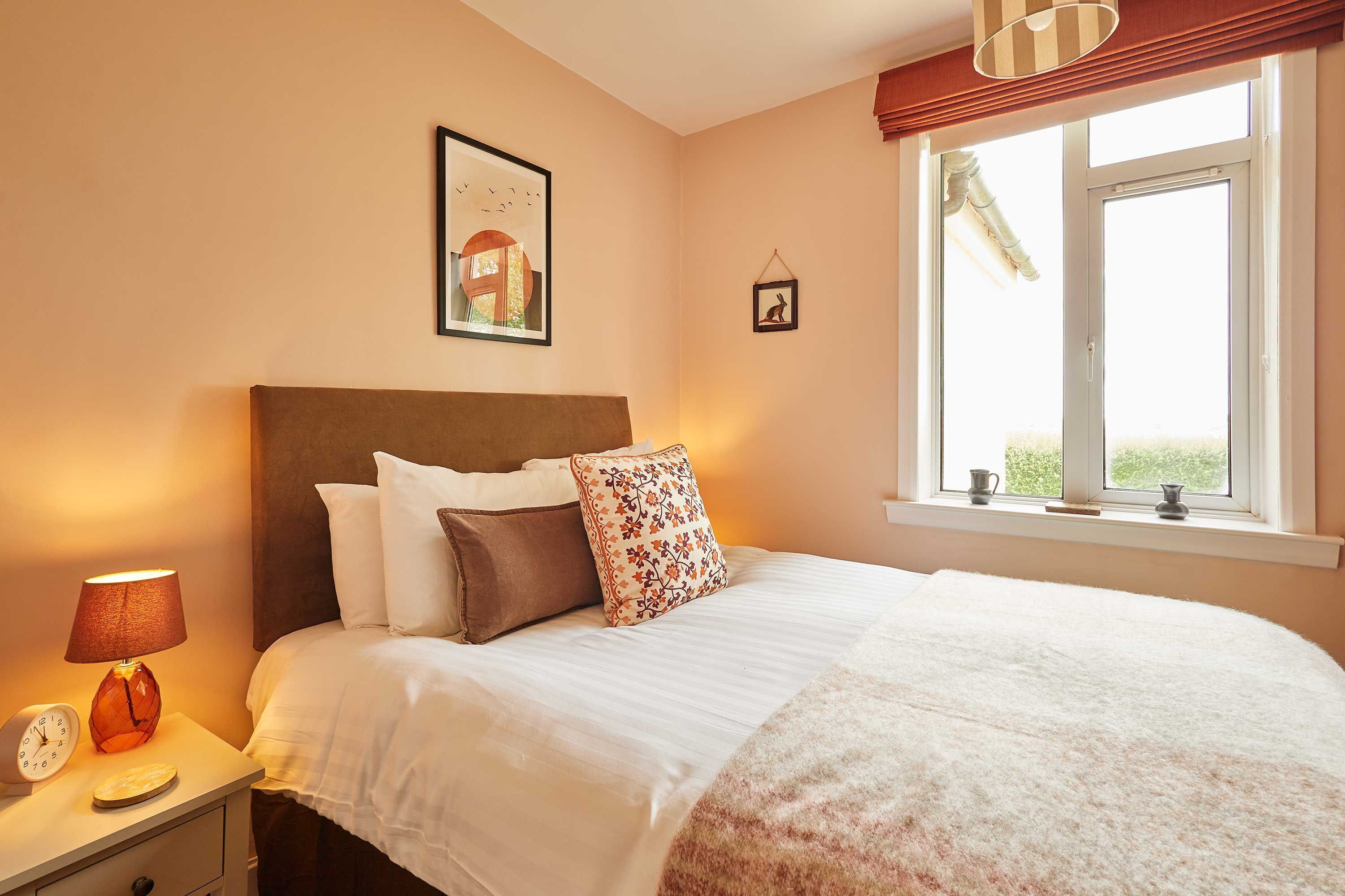 A bedroom with a bed, a nightstand, a lamp, a framed picture, and a window with a view.