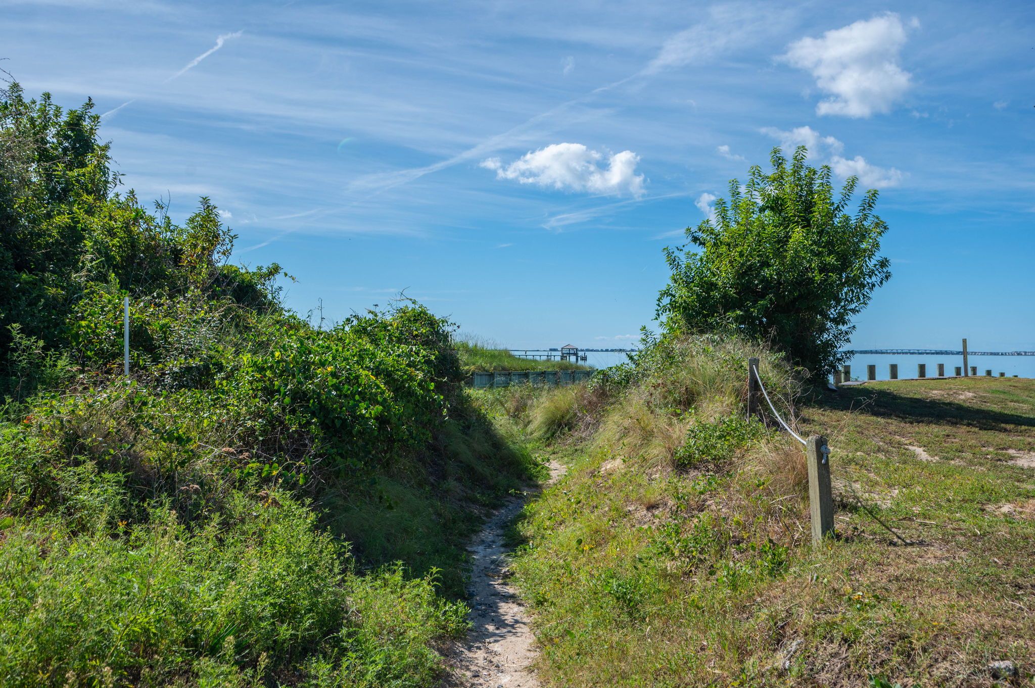 A narrow dirt path leading to a wooden structure with a view of a body of water and a distant bridge.