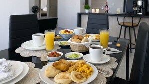 En frukostuppdukning med tallrikar, koppar och ett matbord i ett modernt rum, med ett kök synligt i bakgrunden.
