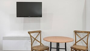 A room with a flat-screen TV mounted on the wall, a white air conditioning unit, and two wicker chairs around a small round table.