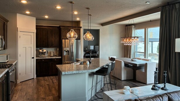 A modern kitchen with a central island, stainless steel appliances, pendant lighting, and a dining area with a view. - Bougie Basecamp: Heated Pool+Hot Tub+Sauna+Trails+Adventure Toy Rental Options (Lehi)