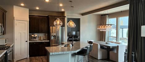 A modern kitchen with a central island, stainless steel appliances, pendant lighting, and a dining area with a view.