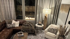 A cozy living room with a leather sofa, a white armchair, a coffee table with a bowl and a book, a lamp, and a view of a fire outside. - Bougie Basecamp: Heated Pool+Hot Tub+Sauna+Trails+Adventure Toy Rental Options (Lehi)