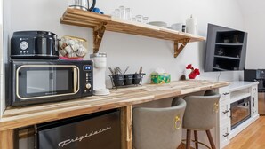 Een compacte keuken met een magnetron, broodrooster en koffiezetapparaat op het aanrecht, een houten plank erboven en een kleine koelkast eronder.