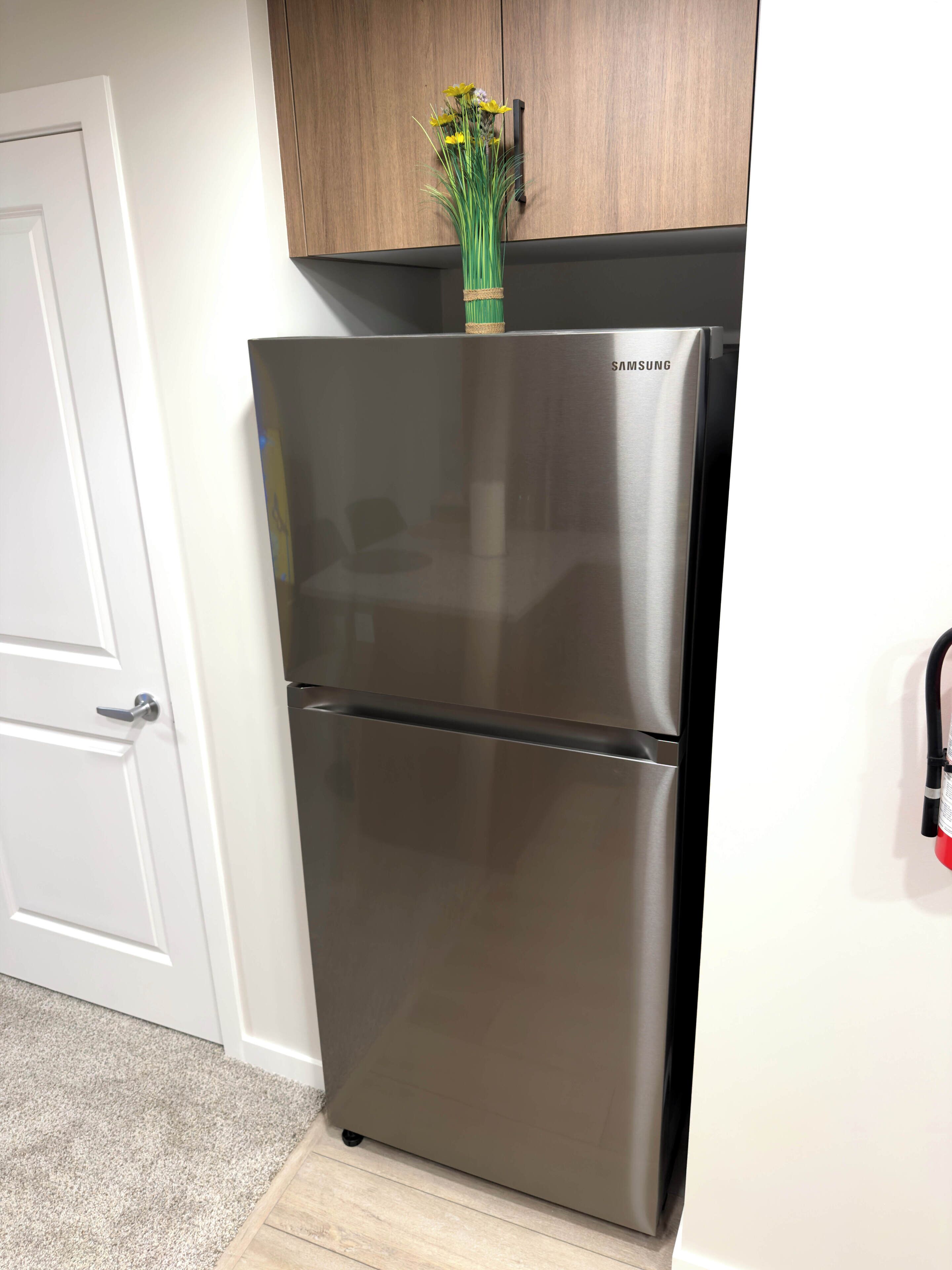 Un refrigerador de acero inoxidable con el logo de Samsung, un gabinete de madera encima y una puerta blanca a la izquierda.