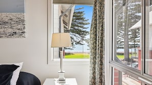 A room with a window, a lamp, a bed, and a view of trees and a building. - 2/16 Flinders Pde, Victor Harbor (Victor Harbor)
