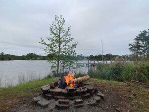 A campfire with logs and a fire pit, surrounded by grass and trees, with a lake and dock in the background.