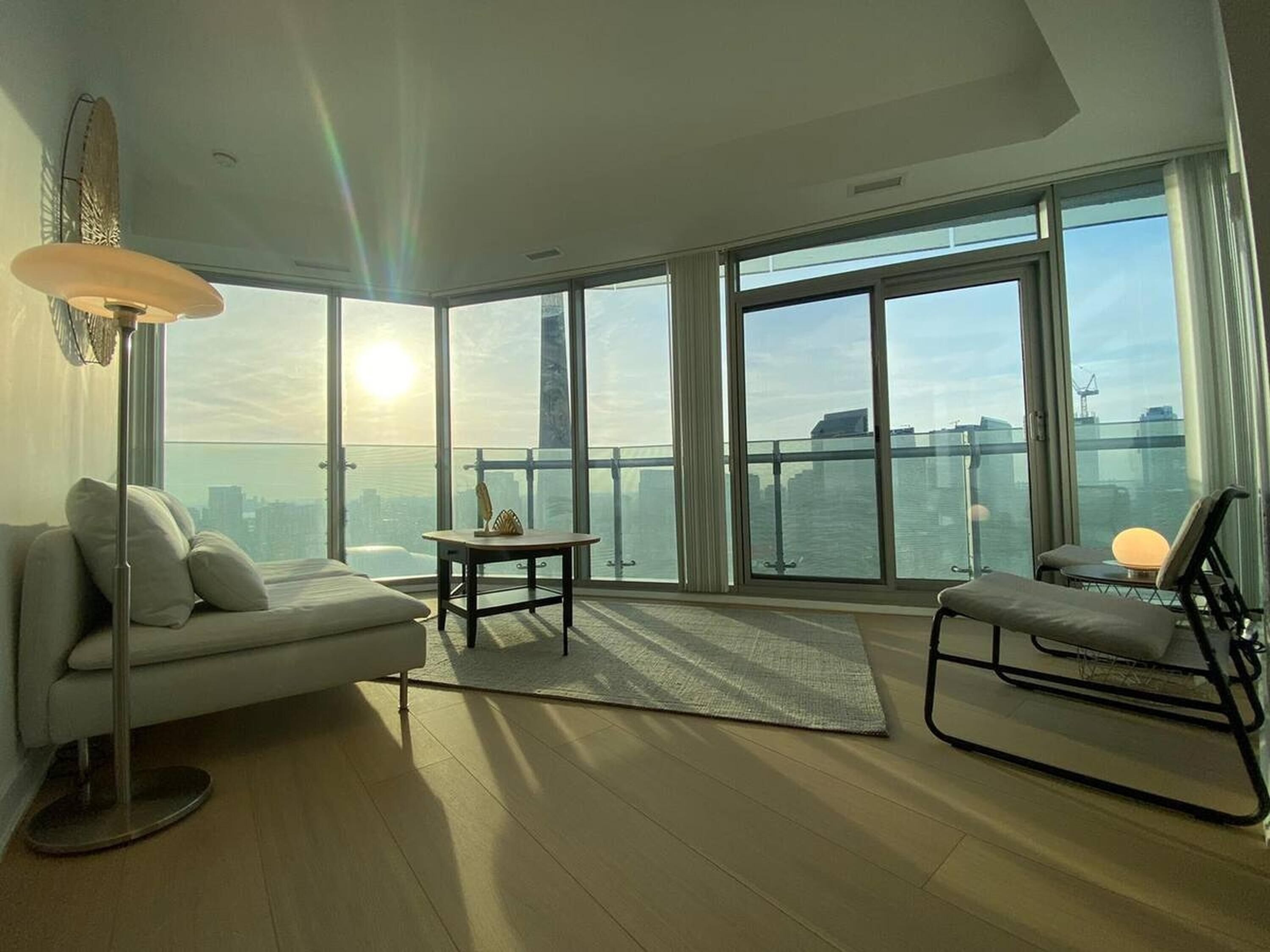 A modern living room with a sofa, a small table, and a chair, large windows with a city view, and a balcony.