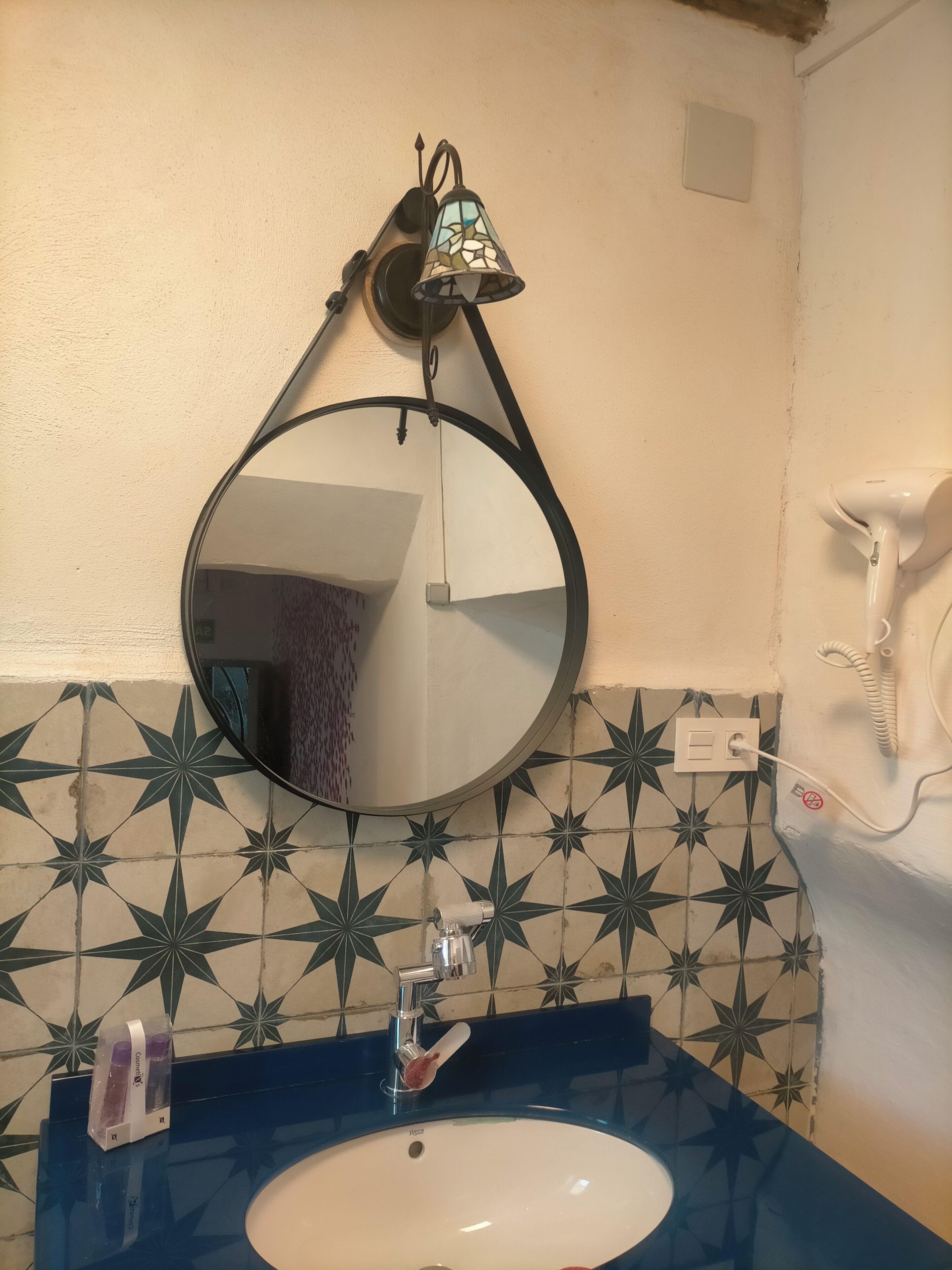 Een badkamer met een ronde spiegel, een wandlamp, een haardroger en een wastafel met een blauw aanrechtblad.