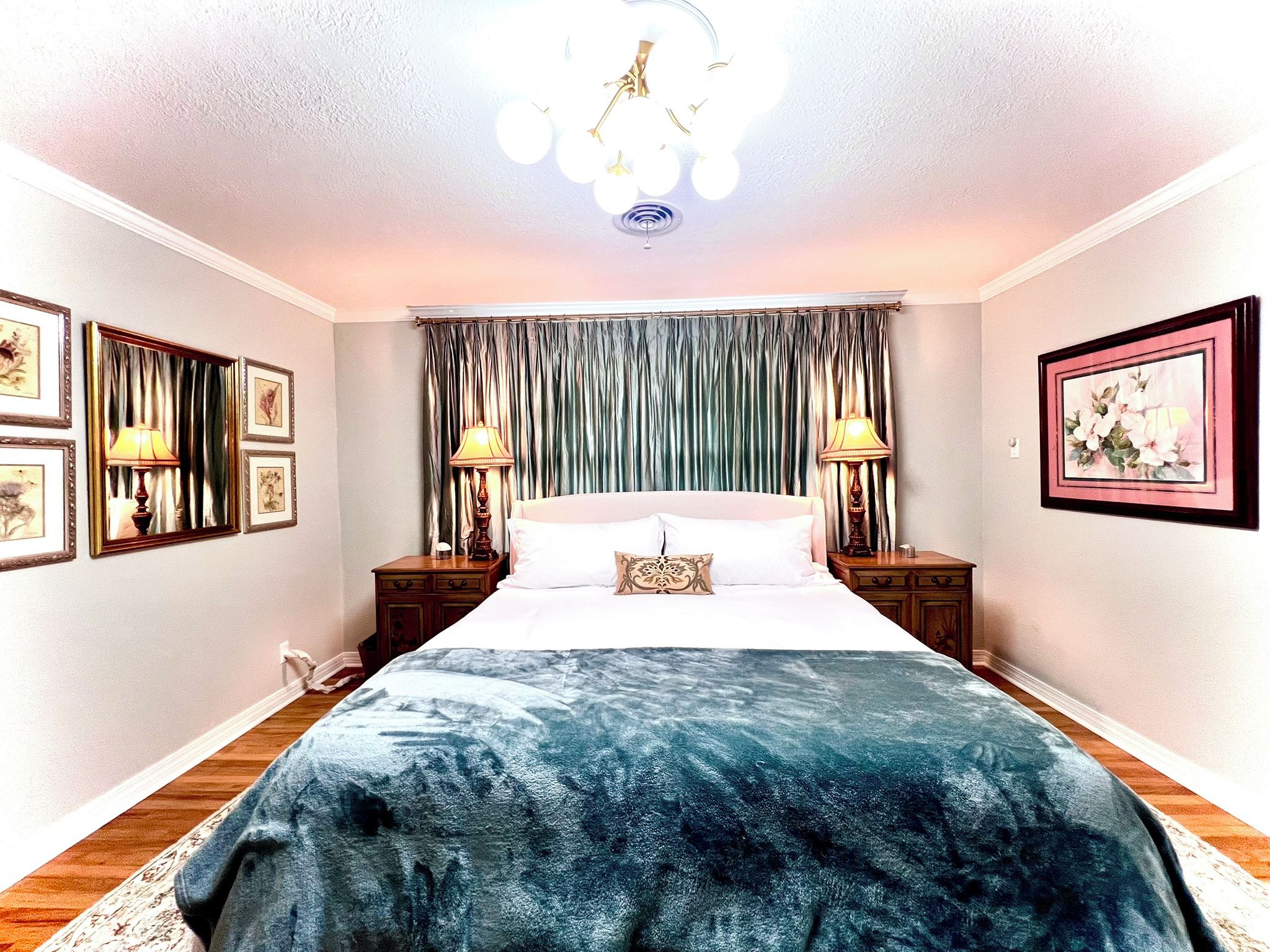 A bedroom with a large bed, two bedside tables with lamps, a chandelier, and framed artwork on the walls.
