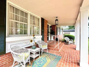 A covered porch with a brick floor, white wicker furniture, and a decorative rug.
